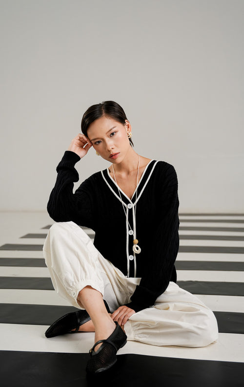 Kardigan wanita Annette Black dengan pola rajut kepang (cable knit), kerah V neck dengan garis putih kontras, dan kancing depan warna putih. 