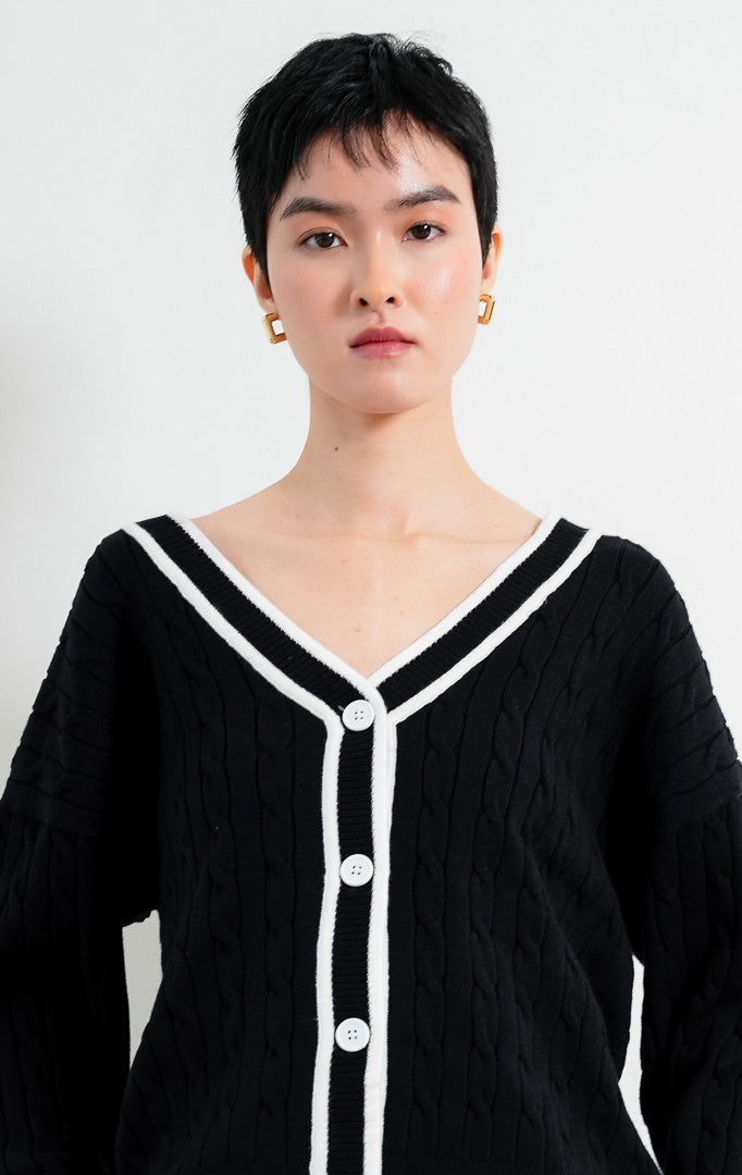 Kardigan rajut wanita lengan panjang warna hitam (black) Annette Cardigan bermotif rajut kepang (cable knit) dengan lis kontras warna putih di leher V (V-neckline) dan bukaan kancing depan. 