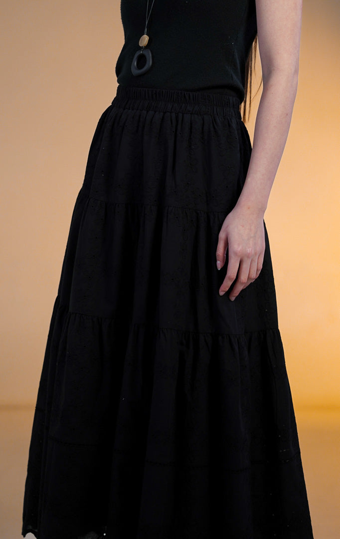 Model wanita mengenakan rok midi hitam model susun (tiered skirt) dengan detail bordir eyelet di bagian bawah, dipadukan dengan tank top hitam dan sandal jepit putih.
