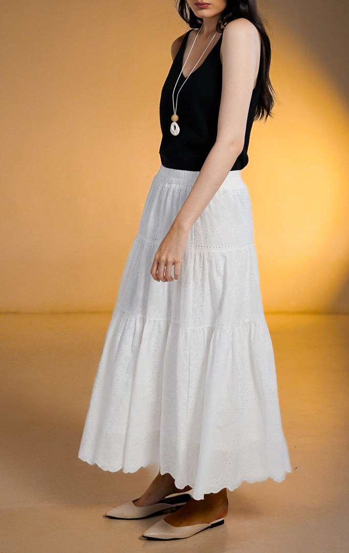 Model wanita mengenakan rok midi putih model susun (tiered skirt) dengan detail bordir eyelet bunga, dipadukan dengan atasan hitam dan sepatu flat putih.