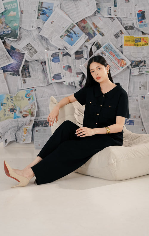 Model wanita mengenakan Arnie Top Black, atasan rajut (knitwear) lengan pendek warna hitam dengan kerah kemeja. Baju memiliki detail kancing emas (gold buttons) di depan dan saku fungsional, dipadukan dengan celana panjang hitam untuk gaya all-black yang elegan.