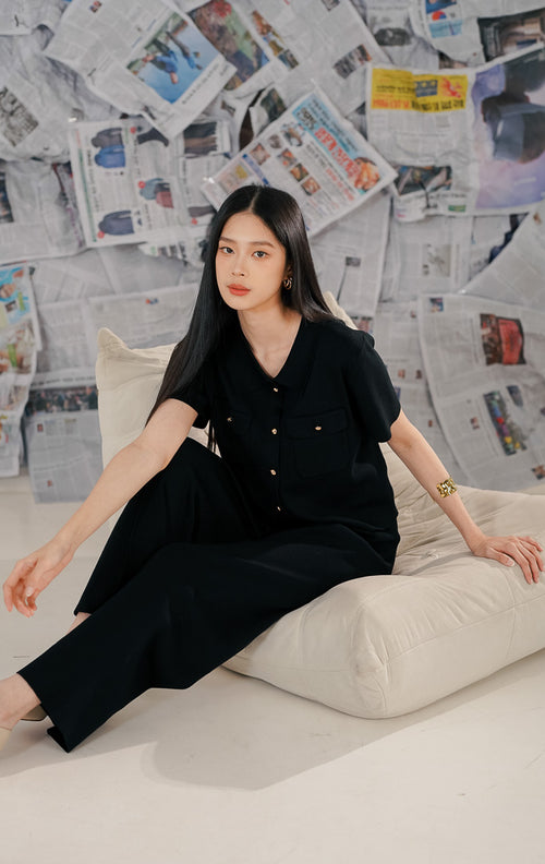 Model wanita mengenakan Arnie Top Black, atasan rajut (knitwear) lengan pendek warna hitam dengan kerah kemeja. Baju memiliki detail kancing emas (gold buttons) di depan dan saku fungsional, dipadukan dengan celana panjang hitam untuk gaya all-black yang elegan.