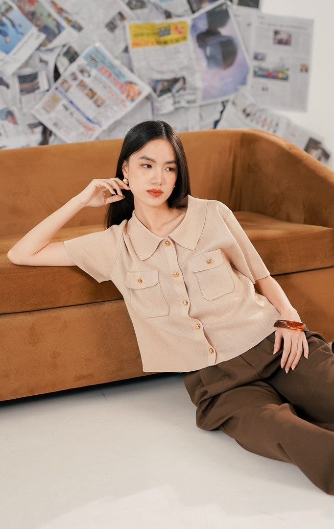 Model wanita mengenakan Arnie Top Brown, sebuah atasan rajut (knit top) lengan pendek warna cokelat muda/beige. Baju memiliki kerah kemeja, dua saku depan, dan kancing emas (gold buttons), dipadukan dengan celana kulot cokelat tua untuk gaya elegan.