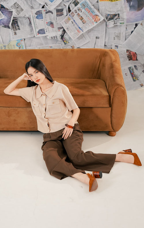 Model wanita mengenakan Arnie Top Brown, sebuah atasan rajut (knit top) lengan pendek warna cokelat muda/beige. Baju memiliki kerah kemeja, dua saku depan, dan kancing emas (gold buttons), dipadukan dengan celana kulot cokelat tua untuk gaya elegan.