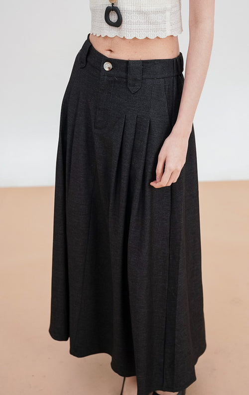 Model mengenakan Ashton Skirt warna black, rok panjang tailored dengan detail lipit (pleats) di bagian depan, ban pinggang rapi dengan satu kancing, dan siluet terstruktur.