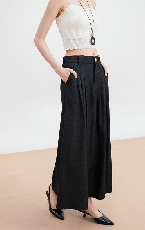 Model mengenakan Ashton Skirt warna black, rok panjang tailored dengan detail lipit (pleats) di bagian depan, ban pinggang rapi dengan satu kancing, dan siluet terstruktur.