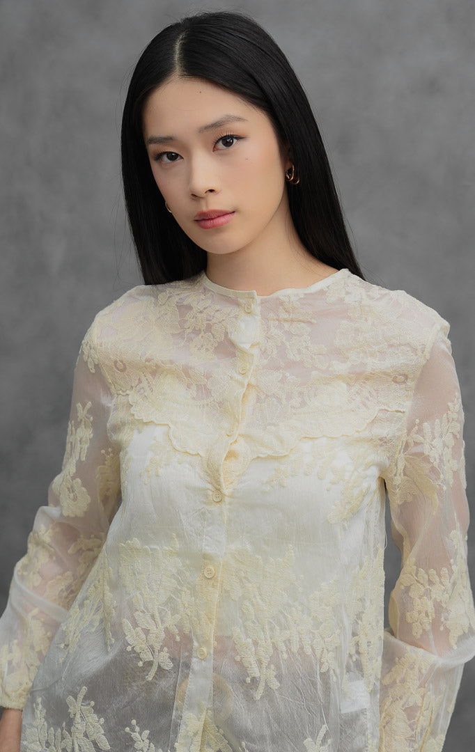 Blus wanita lengan panjang warna krem muda (ivory) Astridia Top berbahan sheer transparan terstruktur dengan detail bordir bunga (floral embroidery) dan bukaan kancing depan.