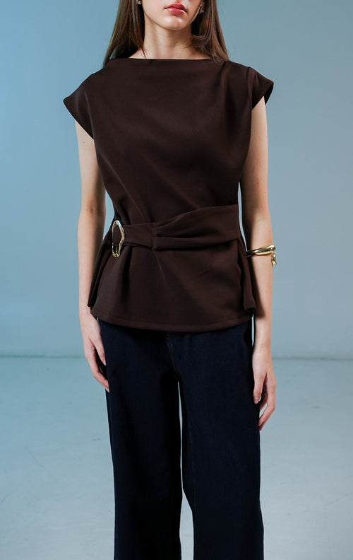 Model wanita mengenakan Carpenter Sleeveless Draped Top warna coklat tua, atasan tanpa lengan berbahan katun poliester dengan siluet drape asimetris dan detail sabuk bergesper di pinggang. 