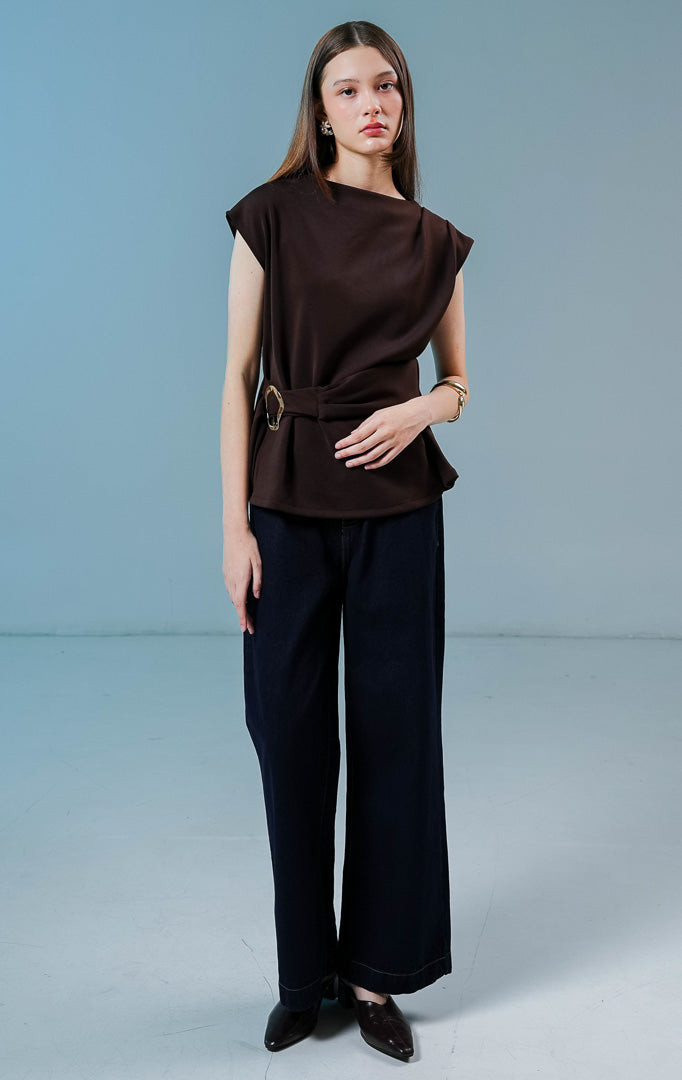Model wanita mengenakan Carpenter Sleeveless Draped Top warna coklat tua, atasan tanpa lengan berbahan katun poliester dengan siluet drape asimetris dan detail sabuk bergesper di pinggang. 