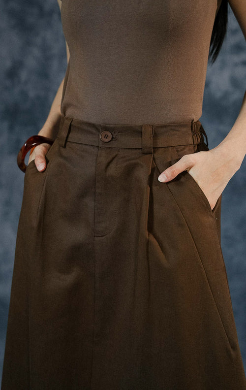 Rok panjang wanita Avella warna dark brown dengan potongan A-line, detail lipit (pleats), kancing depan, dan karet elastis di bagian pinggang belakang.