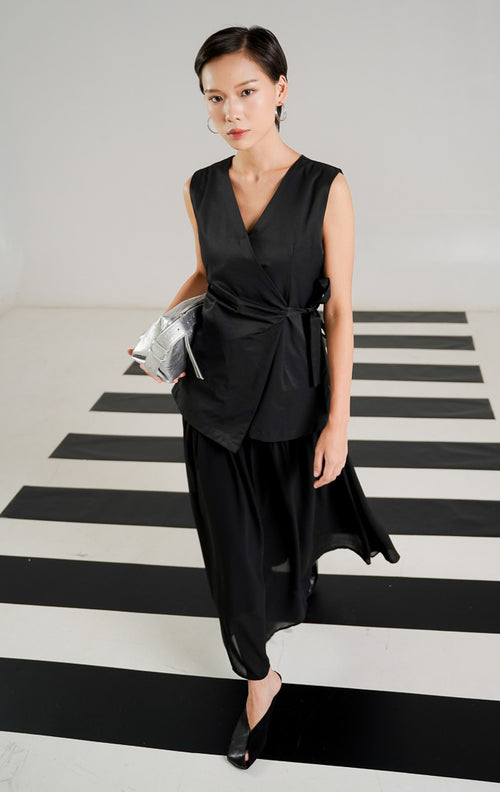 Gaun tanpa lengan Barenia Dress Black dengan model wrap asimetris, detail tali ikat di pinggang, dan kerah V neck dari This Is April. 