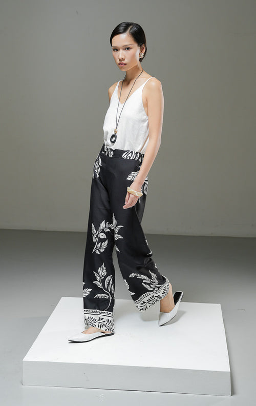 Celana panjang wanita Beatrice Pants warna hitam dengan motif botanical daun dan bunga putih, potongan high waist straight leg, dan ban pinggang rapi.