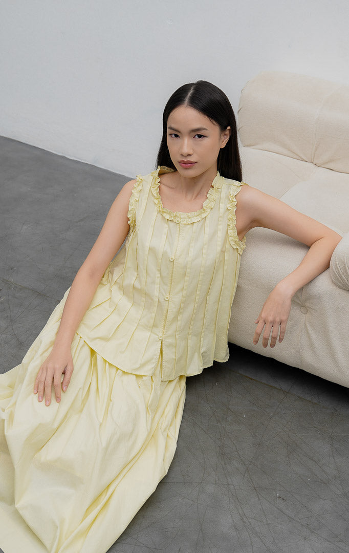 Atasan tanpa lengan (sleeveless) wanita warna kuning lembut (butter yellow) Belin Top berbahan katun (crisp cotton) dengan aksen kerut (ruffle trims), detail lipitan (pintuck detailing), dan bukaan kancing depan.