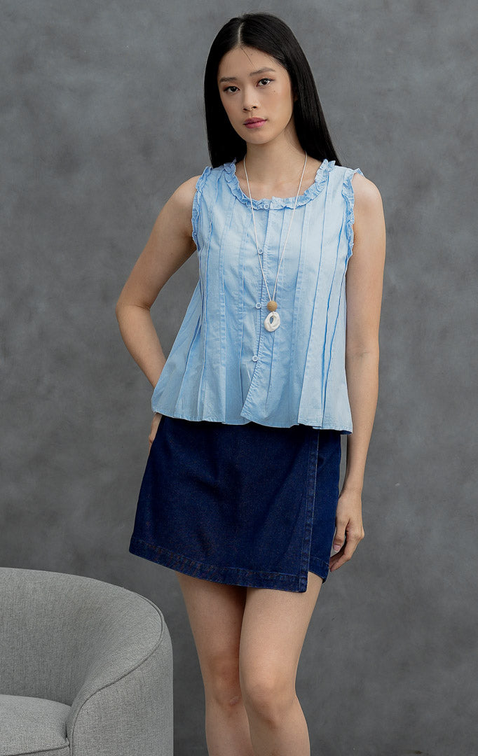 Atasan tanpa lengan (sleeveless) wanita warna biru muda (light blue) Belin Top berbahan katun (crisp cotton) dengan aksen kerut (ruffle trims), detail lipitan (pintuck detailing), dan bukaan kancing depan.