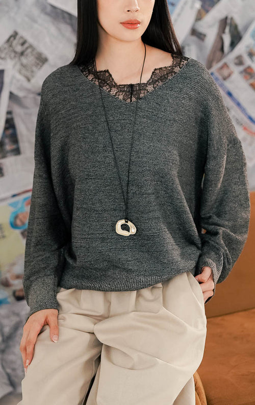 Model wanita mengenakan Bijou Sweater Dark Grey, sebuah atasan rajut lengan panjang warna abu-abu tua. Sweater memiliki potongan leher V-neck dengan detail renda (lace) hitam yang feminin di bagian dada. Dipadukan dengan celana kulot beige longgar untuk tampilan kasual yang nyaman.