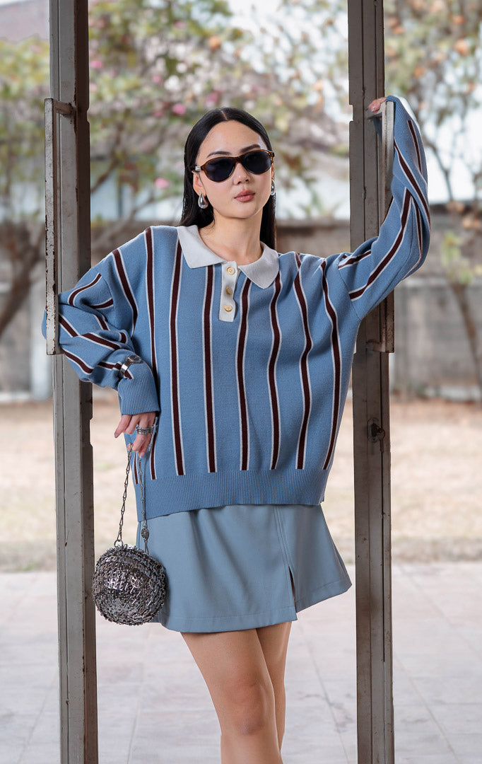 Sweater rajut wanita lengan panjang warna biru muda dengan detail garis vertikal kontras (light blue pinstripe polo knit sweater), kerah polo putih kontras (contrast white polo collar), dan penutup kancing depan (front-button closure).