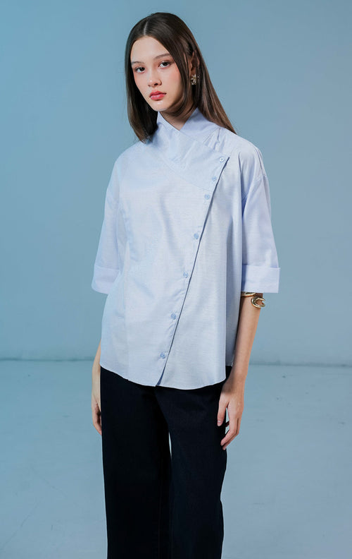 Model wanita mengenakan Brielle Buttons High Neck Top warna biru muda, blouse katun berpotongan elegan dengan kerah tinggi dan detail kancing asimetris di bagian depan. 