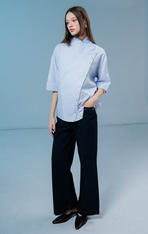 Model wanita mengenakan Brielle Buttons High Neck Top warna biru muda, blouse katun berpotongan elegan dengan kerah tinggi dan detail kancing asimetris di bagian depan. 