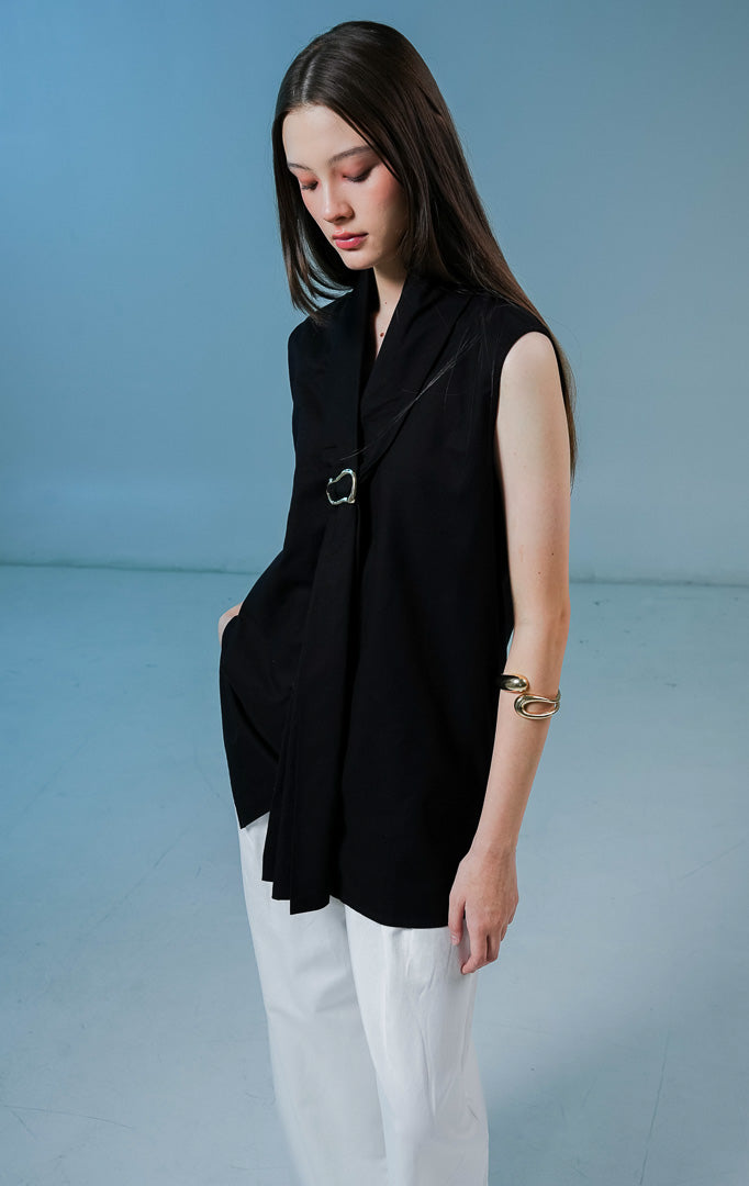 Model wanita mengenakan Fayola Shawl Collar Top warna hitam, atasan tanpa lengan berbahan katun dengan siluet drape yang elegan dan aksen gesper metalik di bagian dada. 