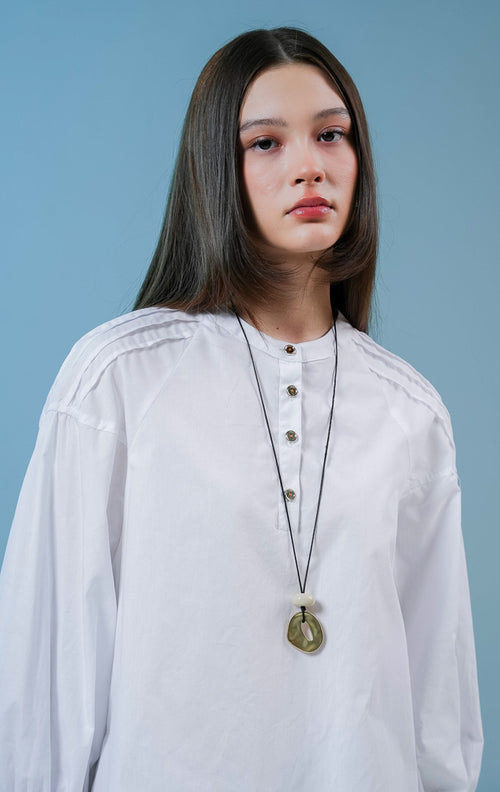 Model wanita mengenakan Cayden Long Sleeve Blouse warna putih, atasan katun berkerah bulat dengan detail lengan panjang beraksen ruffle dan kancing emas di bagian depan. 