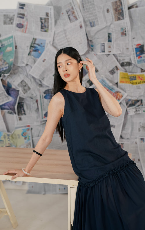 Model wanita mengenakan Brandy Dress Navy, sebuah dress panjang (maxi dress) tanpa lengan warna biru dongker. Dress memiliki potongan leher bulat dan detail ruffle (kerut) pada bagian pinggul (drop waist), memberikan siluet yang flowy dan elegan.