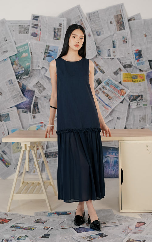 Model wanita mengenakan Brandy Dress Navy, sebuah dress panjang (maxi dress) tanpa lengan warna biru dongker. Dress memiliki potongan leher bulat dan detail ruffle (kerut) pada bagian pinggul (drop waist), memberikan siluet yang flowy dan elegan.