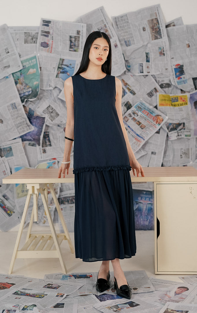 Model wanita mengenakan Brandy Dress Navy, sebuah dress panjang (maxi dress) tanpa lengan warna biru dongker. Dress memiliki potongan leher bulat dan detail ruffle (kerut) pada bagian pinggul (drop waist), memberikan siluet yang flowy dan elegan.
