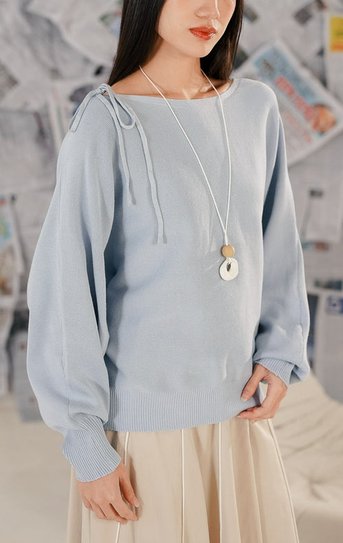 Model wanita mengenakan Brigitte Sweater Blue, sebuah atasan rajut lengan panjang warna biru muda (baby blue). Sweater memiliki potongan leher boat neck dan detail pita dekoratif pada satu sisi bahu. Dipadukan dengan rok midi warna beige dan kalung panjang.