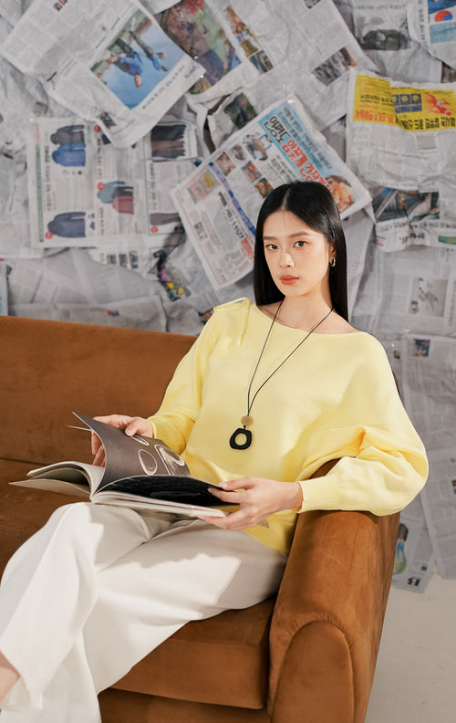 Model wanita mengenakan Brigitte Sweater Yellow, sebuah atasan rajut lengan panjang warna kuning pastel. Sweater memiliki potongan leher melebar (boat neck) dan detail pita dekoratif di bagian bahu/lengan, dipadukan dengan celana panjang putih.