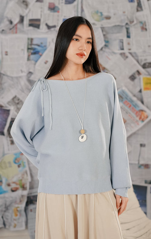 Model wanita mengenakan Brigitte Sweater Blue, sebuah atasan rajut lengan panjang warna biru muda (baby blue). Sweater memiliki potongan leher boat neck dan detail pita dekoratif pada satu sisi bahu. Dipadukan dengan rok midi warna beige dan kalung panjang.
