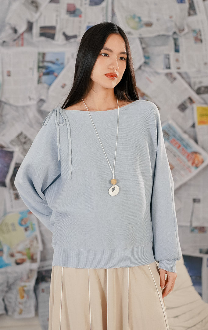 Model wanita mengenakan Brigitte Sweater Blue, sebuah atasan rajut lengan panjang warna biru muda (baby blue). Sweater memiliki potongan leher boat neck dan detail pita dekoratif pada satu sisi bahu. Dipadukan dengan rok midi warna beige dan kalung panjang.