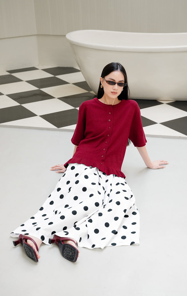 Atasan rajut wanita lengan pendek Brinley Top warna maroon dengan kerah bulat, kancing depan, dan aksen rumbai ruffle hem yang feminin. 