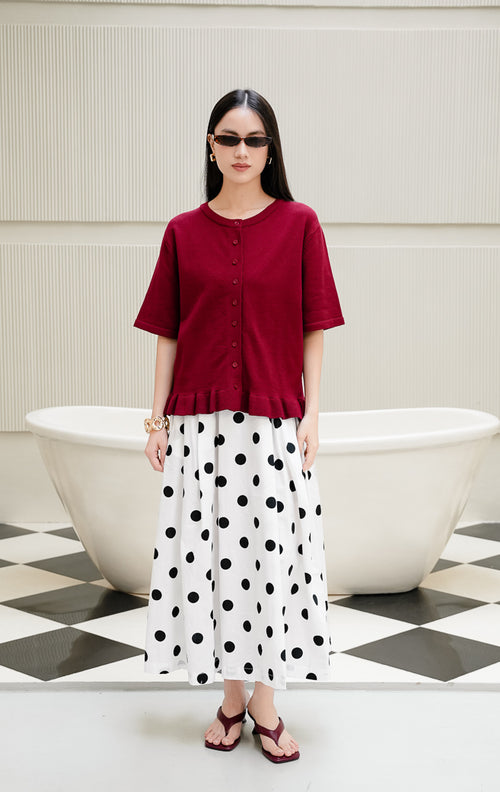 Atasan rajut wanita lengan pendek Brinley Top warna maroon dengan kerah bulat, kancing depan, dan aksen rumbai ruffle hem yang feminin. 