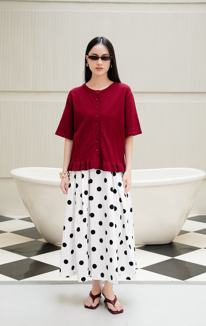 Atasan rajut wanita lengan pendek Brinley Top warna maroon dengan kerah bulat, kancing depan, dan aksen rumbai ruffle hem yang feminin. 