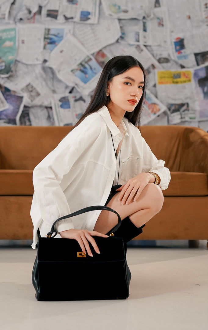 Model wanita mengenakan Bryan Top White, kemeja putih bahan katun dengan potongan lengan panjang yang longgar (relaxed fit). Kemeja memiliki kerah kemeja klasik, kancing depan, dan saku dada, dipadukan dengan rok mini gelap dan sepatu boots tinggi hitam.