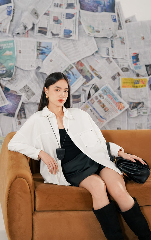 Model wanita mengenakan Bryan Top White, kemeja putih bahan katun dengan potongan lengan panjang yang longgar (relaxed fit). Kemeja memiliki kerah kemeja klasik, kancing depan, dan saku dada, dipadukan dengan rok mini gelap dan sepatu boots tinggi hitam.