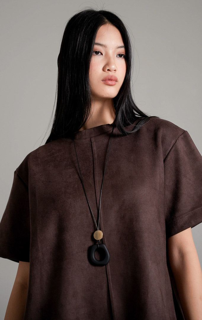 Callidora Suede Top lengan pendek warna cokelat tua bersiluet longgar, dipadukan dengan celana panjang gading, kalung panjang, dan sepatu mules hitam.