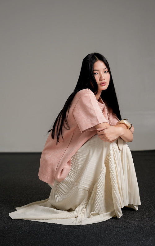 Callidora Suede Top lengan pendek warna pink pastel bersiluet longgar, dipadukan dengan rok panjang lipit warna krem dan kalung liontin.