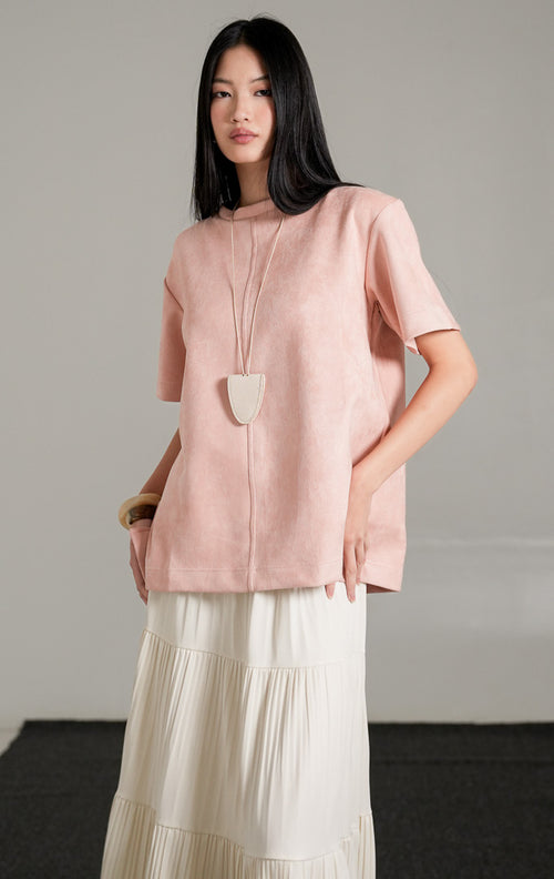 Callidora Suede Top lengan pendek warna pink pastel bersiluet longgar, dipadukan dengan rok panjang lipit warna krem dan kalung liontin.