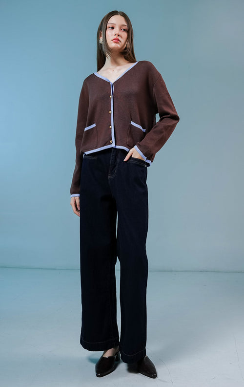 Model wanita mengenakan Darien V-Neck Cardigan warna coklat tua, luaran cardigan rajut wanita dengan detail garis tepi kontras berwarna biru muda, saku depan, dan kancing. 