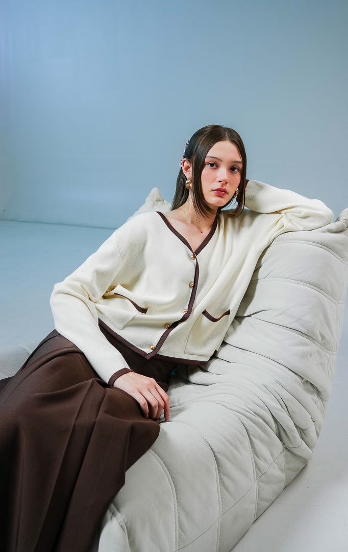 Model wanita mengenakan Darien V-Neck Cardigan warna ivory (krem), luaran rajut wanita dengan detail garis tepi kontras berwarna coklat tua, saku depan, dan kancing. 