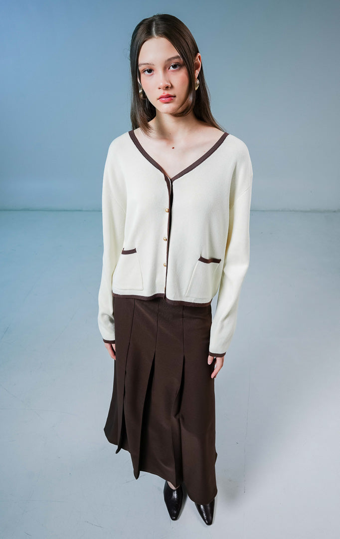 Model wanita mengenakan Darien V-Neck Cardigan warna ivory (krem), luaran rajut wanita dengan detail garis tepi kontras berwarna coklat tua, saku depan, dan kancing. 