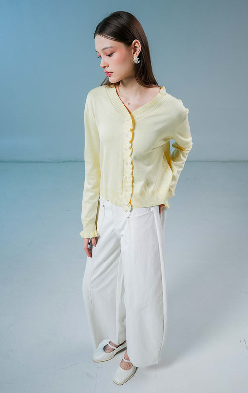 Model wanita mengenakan Graciella Ruffle Knit Cardigan warna butter yellow, luaran rajut wanita berkerah V dengan detail kerutan (ruffles) yang feminin dan kancing mutiara depan. 