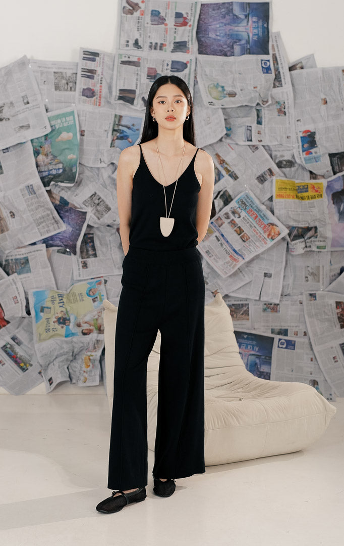 Model wanita mengenakan Carrie Pants Black, celana panjang rajut warna hitam dengan potongan kaki lebar (wide leg). Celana memiliki detail garis jahitan vertikal di bagian depan kaki dan bahan yang jatuh (flowy), dipadukan dengan sepatu flat hitam.