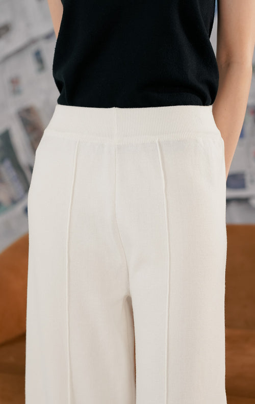 Model wanita mengenakan Carrie Pants White, celana panjang rajut (knit trousers) warna putih tulang dengan potongan kaki lebar (wide leg). Celana memiliki detail garis jahitan vertikal di bagian depan kaki dan pinggang karet, dipadukan dengan sepatu flat hitam berpayet.