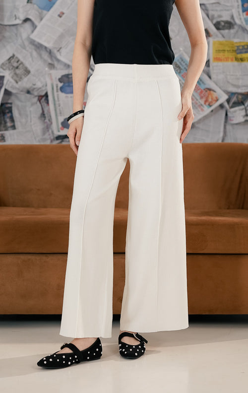 Model wanita mengenakan Carrie Pants White, celana panjang rajut (knit trousers) warna putih tulang dengan potongan kaki lebar (wide leg). Celana memiliki detail garis jahitan vertikal di bagian depan kaki dan pinggang karet, dipadukan dengan sepatu flat hitam berpayet.