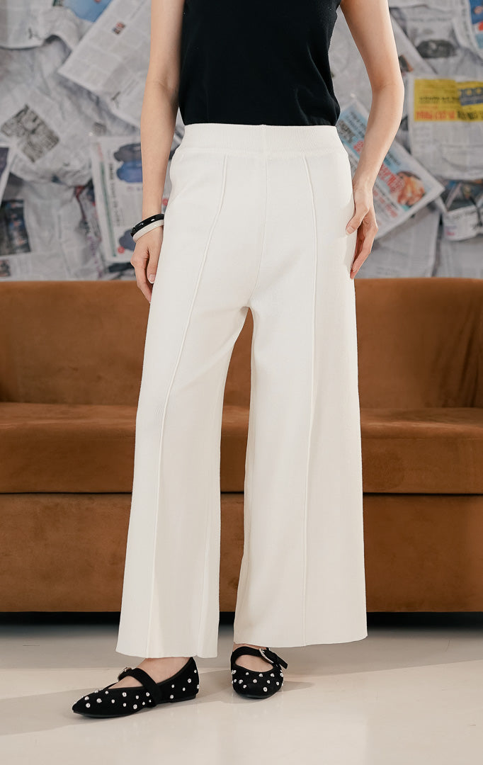 Model wanita mengenakan Carrie Pants White, celana panjang rajut (knit trousers) warna putih tulang dengan potongan kaki lebar (wide leg). Celana memiliki detail garis jahitan vertikal di bagian depan kaki dan pinggang karet, dipadukan dengan sepatu flat hitam berpayet.