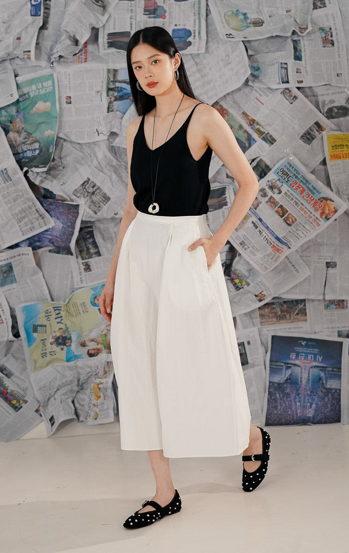 Model wanita mengenakan Casette Skirt White, rok midi model A-line warna putih dari bahan katun. Rok memiliki saku samping tersembunyi dan tampilan minimalis, dipadukan dengan atasan hitam dan sepatu flat untuk gaya kasual.