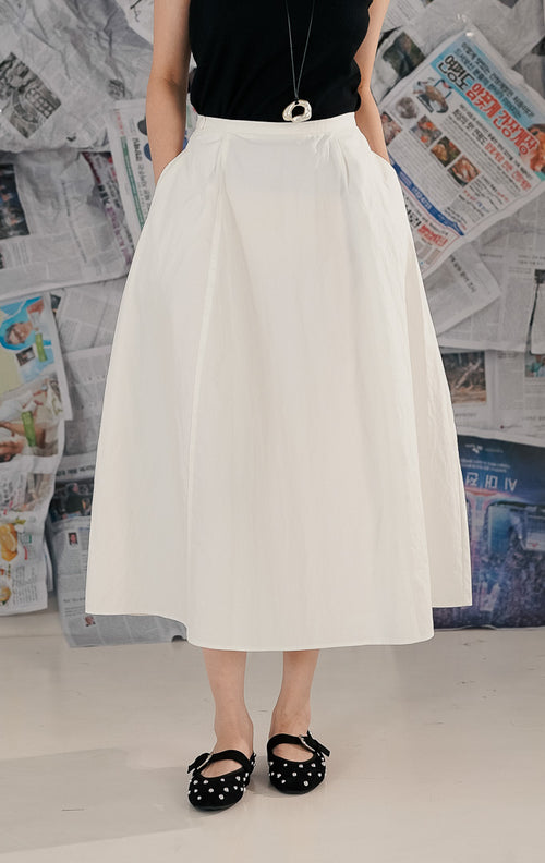 Model wanita mengenakan Casette Skirt White, rok midi model A-line warna putih dari bahan katun. Rok memiliki saku samping tersembunyi dan tampilan minimalis, dipadukan dengan atasan hitam dan sepatu flat untuk gaya kasual.