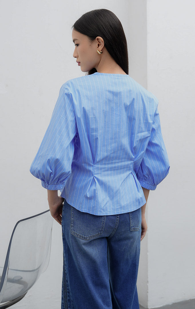 Blus wanita motif garis warna biru (blue) Castell Top berbahan cotton-polyester blend dengan kerah V, aksen kerutan di pinggang samping (side-waist ruching), kancing dekoratif, dan lengan 3/4 bermodel balon.